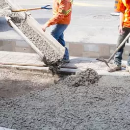 Workers pouring concrete onto a sidewalk from a chute, with tools nearby.