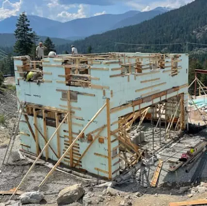Workers assembling a two-story wooden structure on a construction site surrounded by trees and mountains.