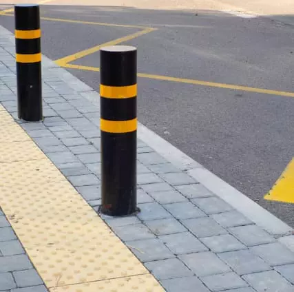 Black bollards with yellow stripes on a tiled sidewalk next to a road with yellow lane markings.