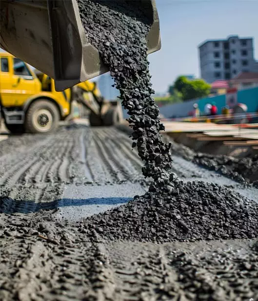 Close-up of a bulldozer pouring wet concrete onto a construction site, with tire tracks visible. A building and a yellow truck are in the background.