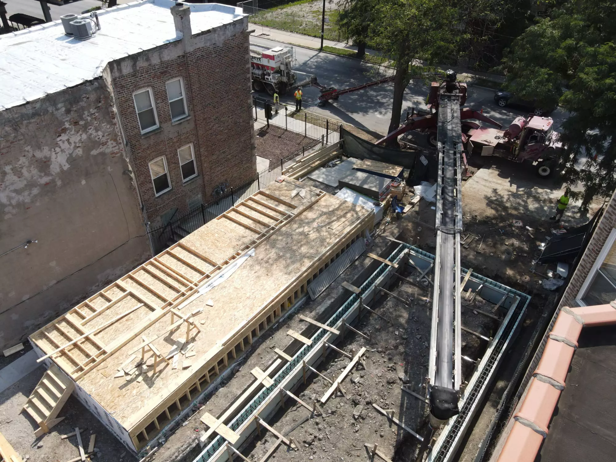 Aerial view of a construction site with workers, a partially built structure with wooden frames, and a crane truck extending its arm. Nearby, a brick building and a tree are visible.