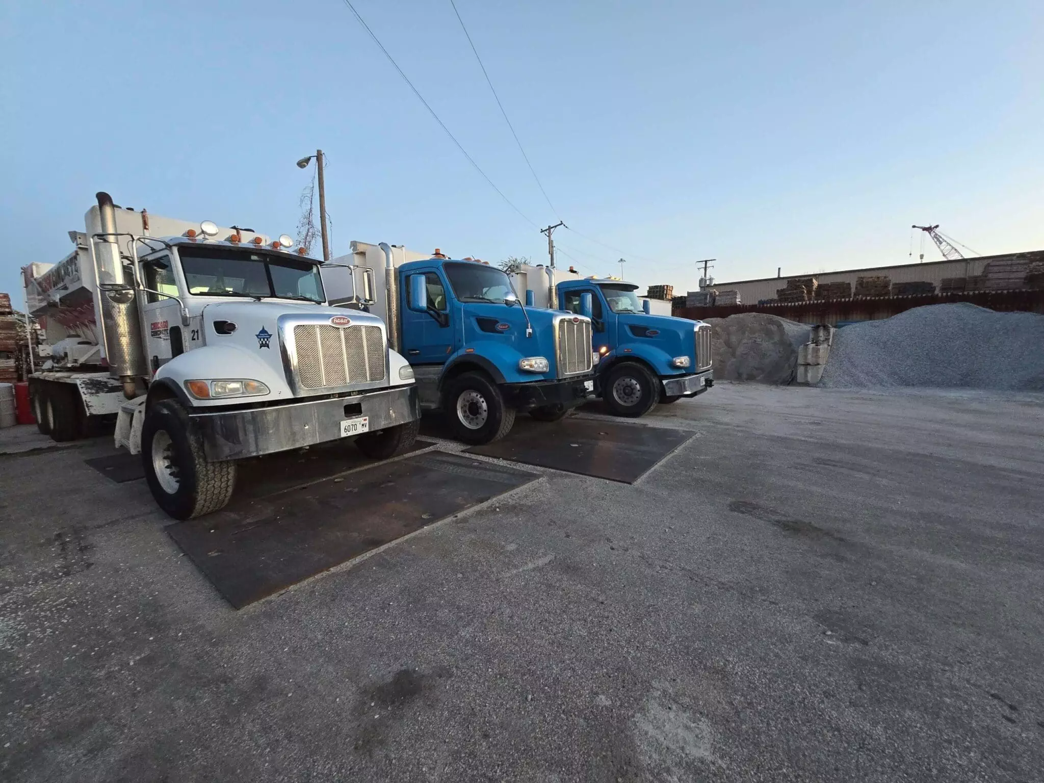 Three parked trucks on an industrial lot with piles of gravel in the background. The sky is clear at dusk.
