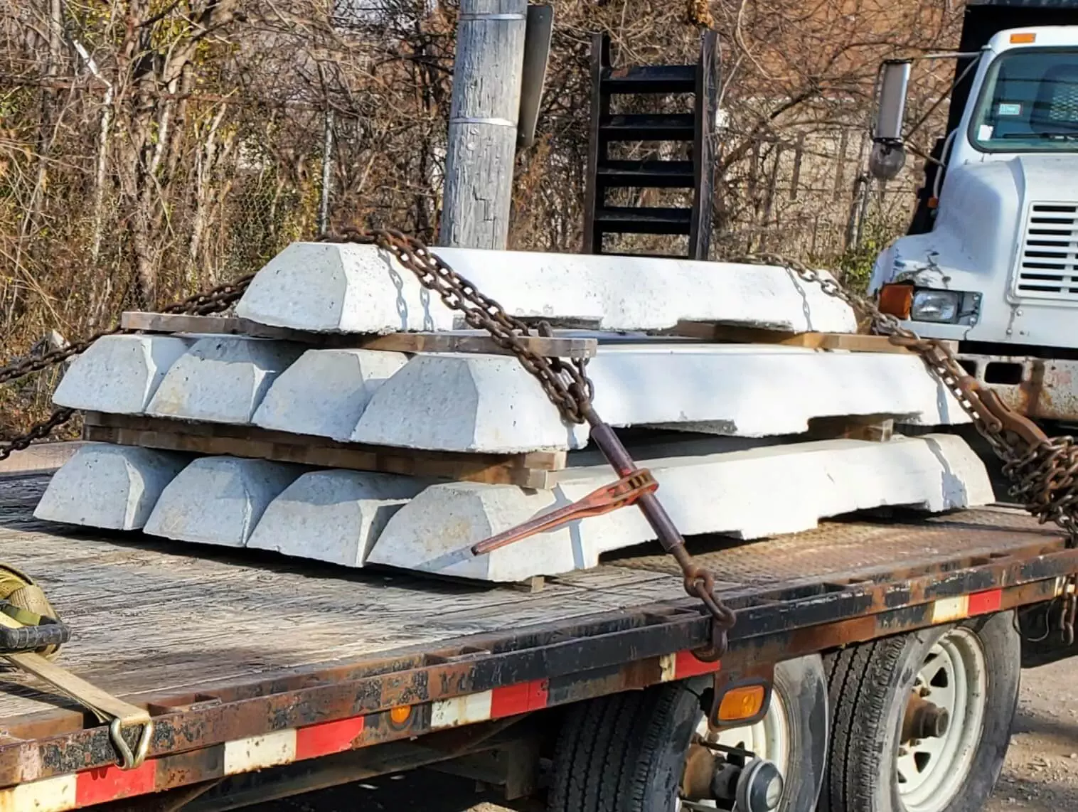 Concrete blocks secured with chains on a flatbed truck outdoors, with a background of trees and another truck.
