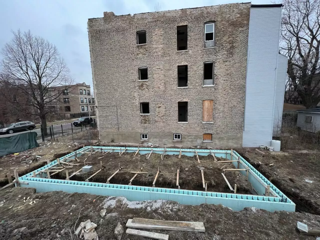 Construction site with an unfinished building foundation in front of an old, four-story brick building with boarded windows. Sparse trees and neighboring buildings are visible in the background.