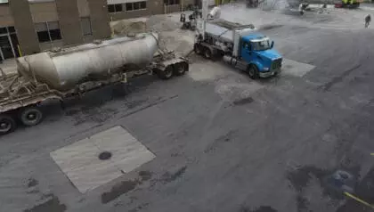 A blue truck is parked next to a tanker trailer in an industrial lot, with buildings and machinery in the background. Workers and vehicles are visible in the distance.